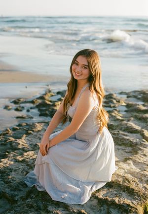 A person in a flowy grey dress sits gracefully on rocky beach terrain at sunset with ocean waves in the background.