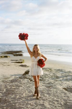 Woman in white dress poses joyfully with red flowers on a rocky beach at sunset.