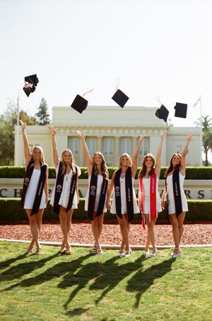Group of graduates in black dresses and stoles toss graduation caps on campus lawn during celebratory photo.