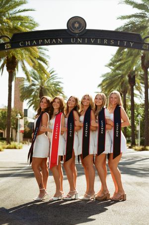 A group of graduates wearing stoles pose together under the Chapman University entrance archway with palm trees.