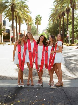 A group in white dresses and red graduation stoles pose together on a palm tree-lined street during a sunny day.
