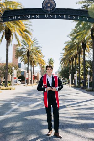 Graduate stands under Chapman University entrance arch with palm trees lining campus pathway on sunny day.