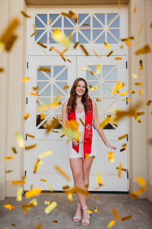 Graduate celebrating achievement with gold confetti falling in bright hallway wearing red cap and gown.