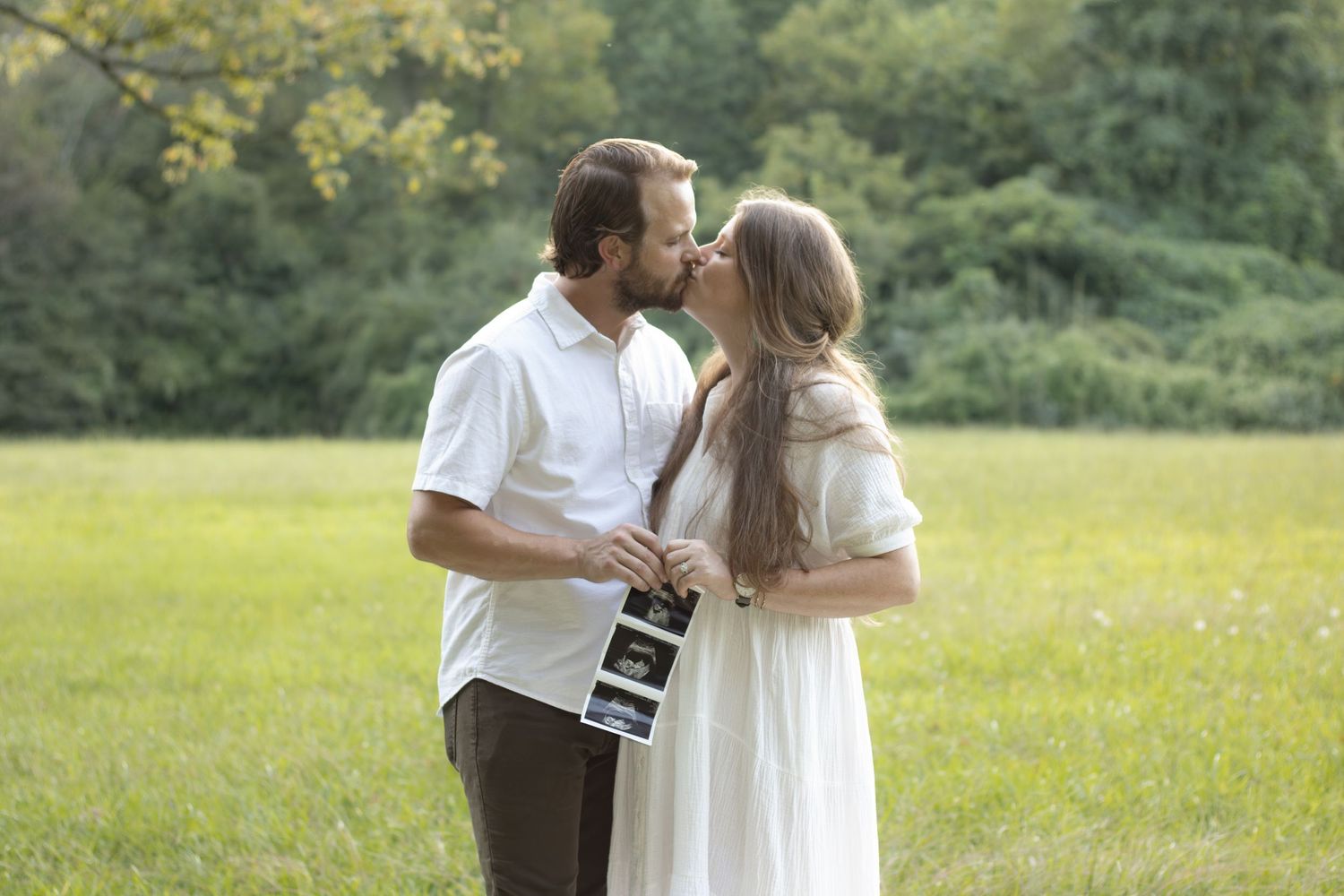Couple sharing intimate moments in a sunny meadow with tall grass and trees in the background.