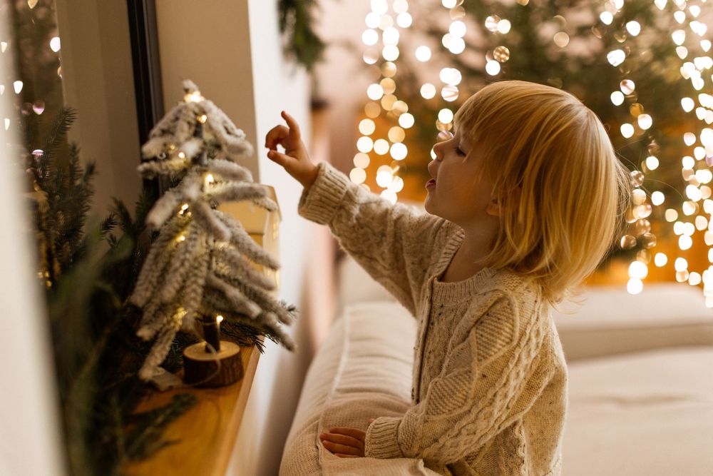 Young blonde child in a cream sweater reaching toward a small decorated Christmas tree with bokeh lights.