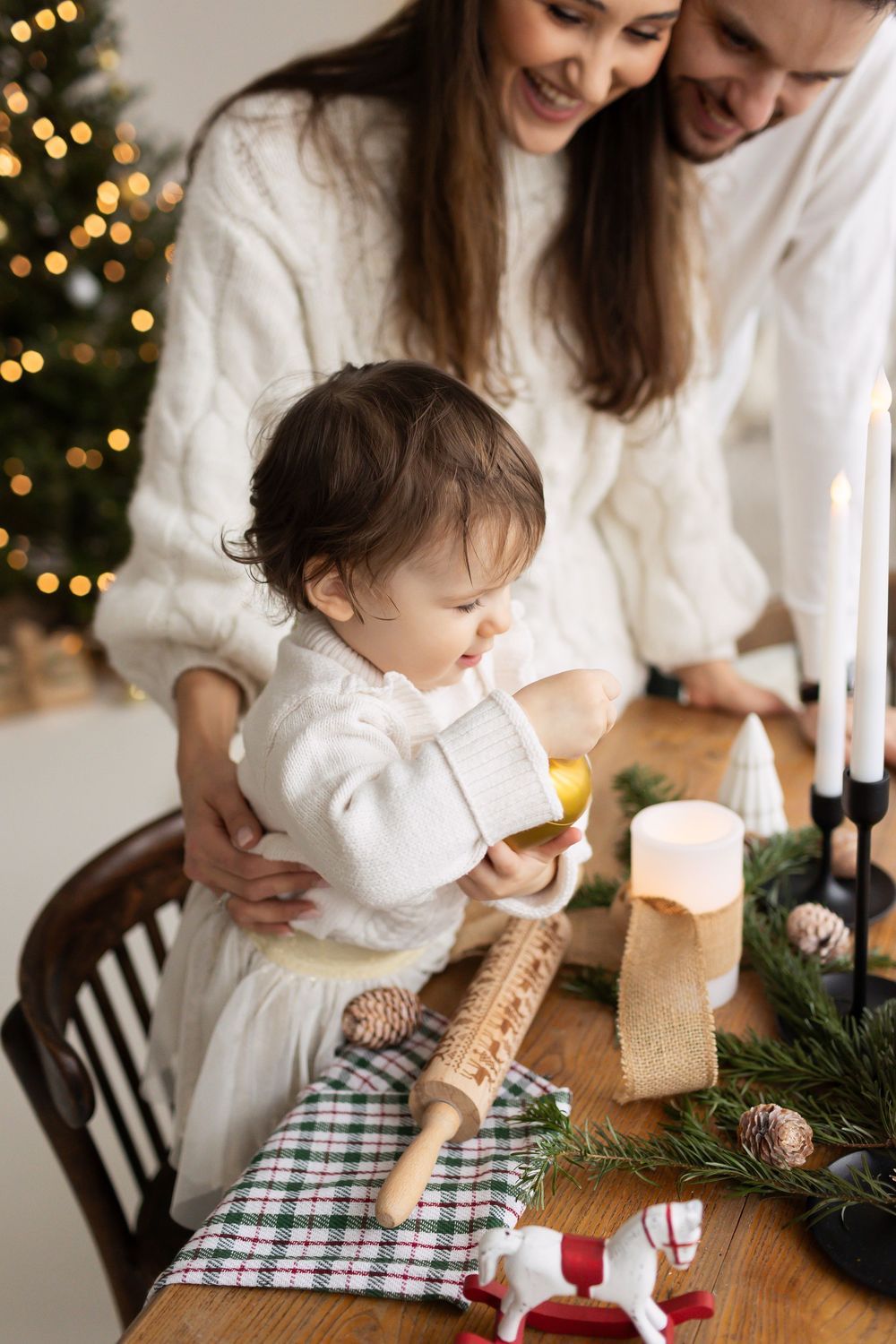 Parents in white sweaters smiling as their baby explores a festive table with candles and Christmas decorations.