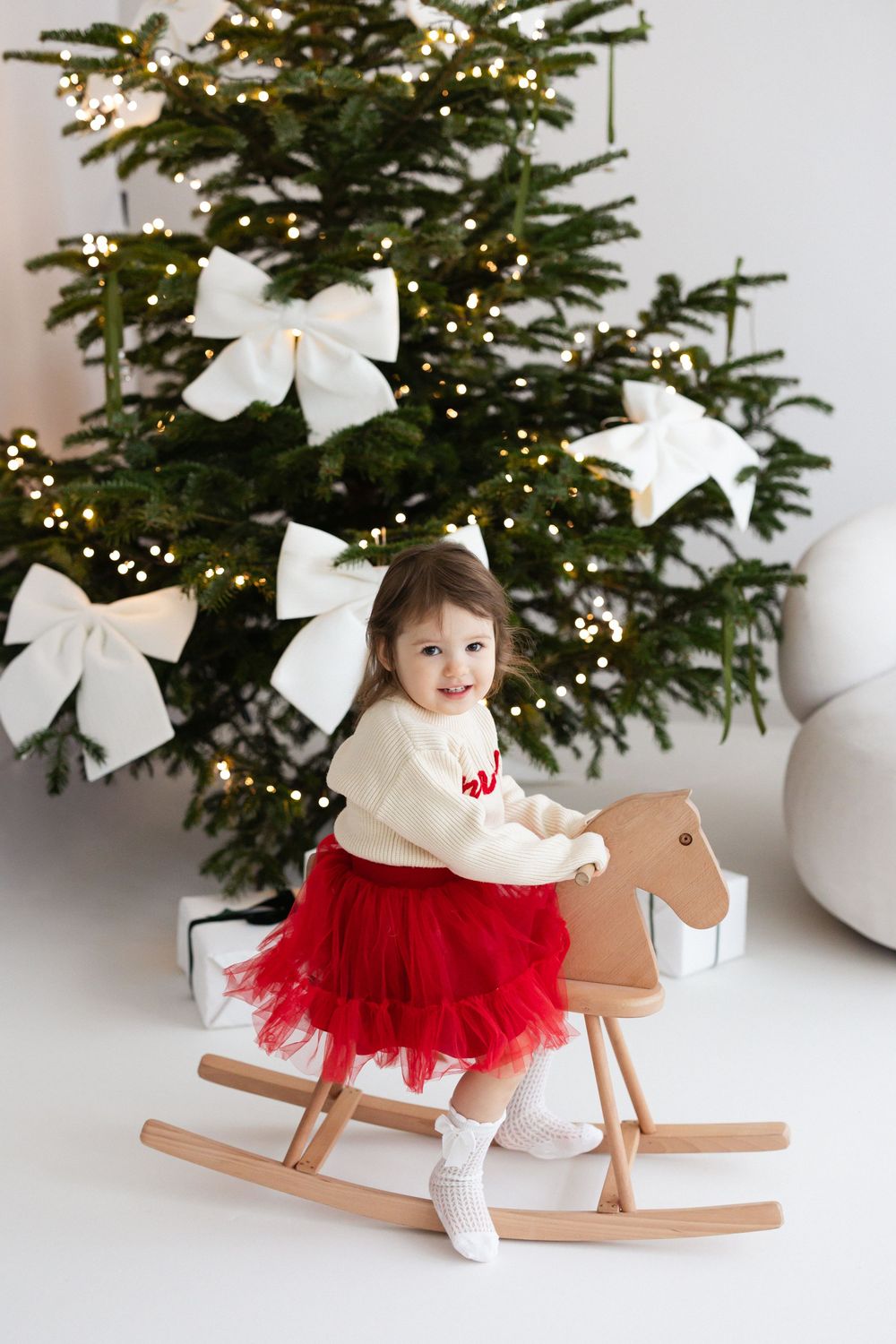 Toddler in a red tulle skirt and cream sweater sits on a wooden rocking horse beside a Christmas tree with white bows and twinkling lights.