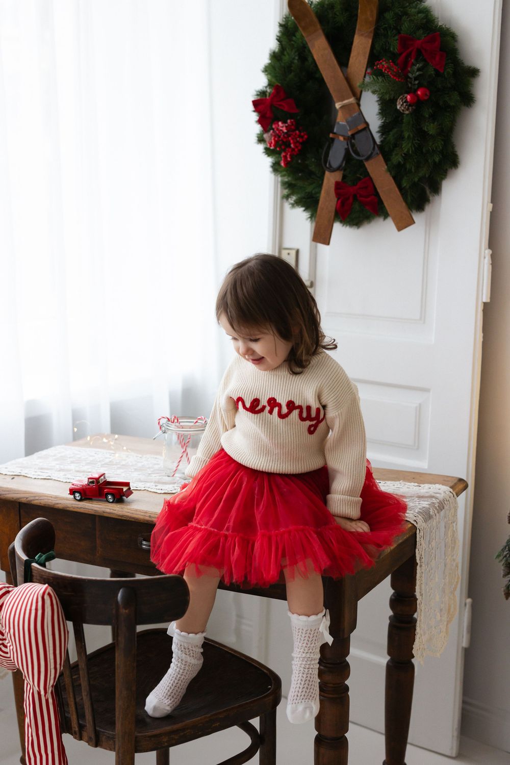Toddler in a cream "merry" sweater and red tulle skirt sits on a wooden desk beside a Christmas wreath with vintage skis.