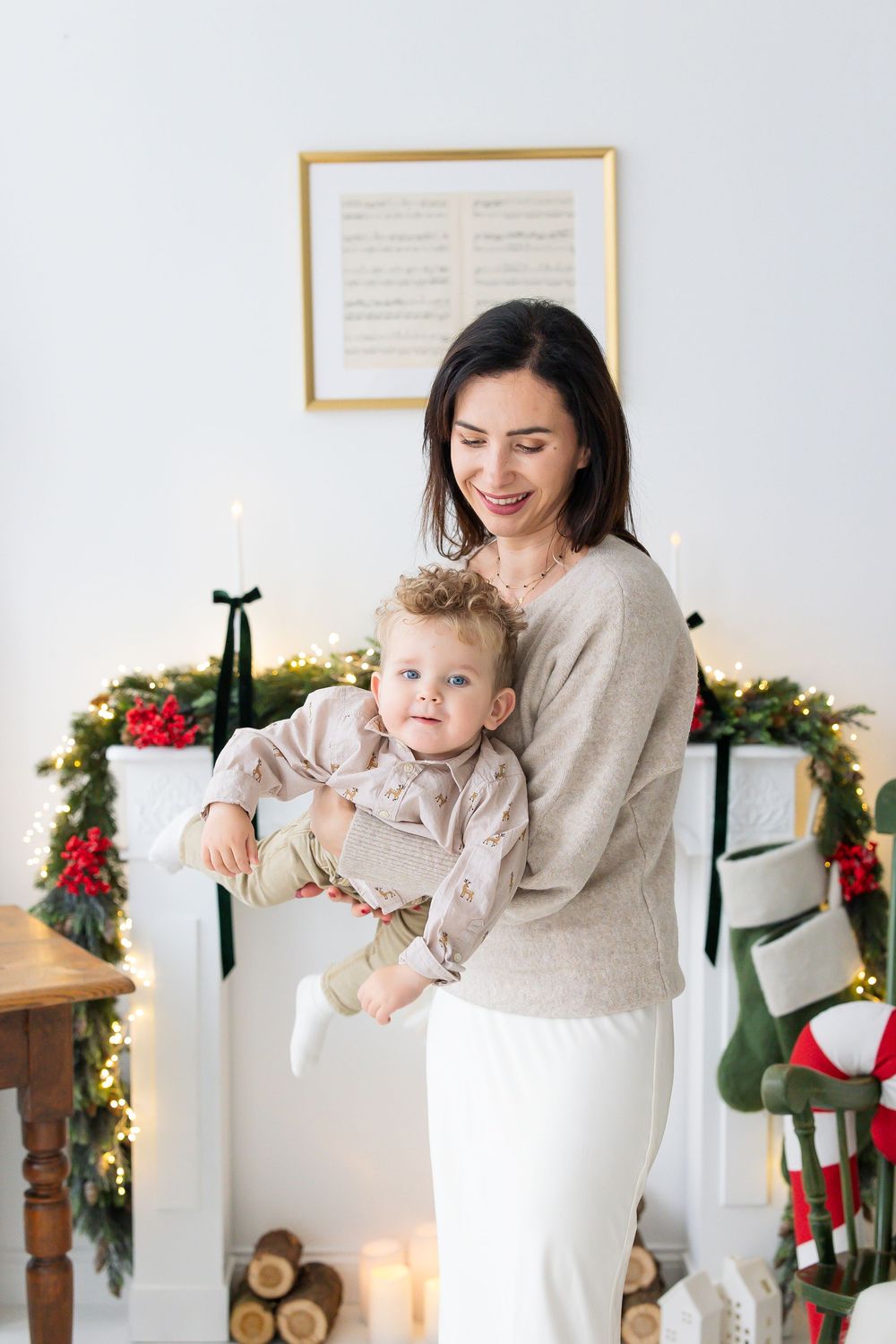 Smiling mother in a cream sweater holds her baby boy beside a white fireplace decorated with a lit Christmas garland and stockings.