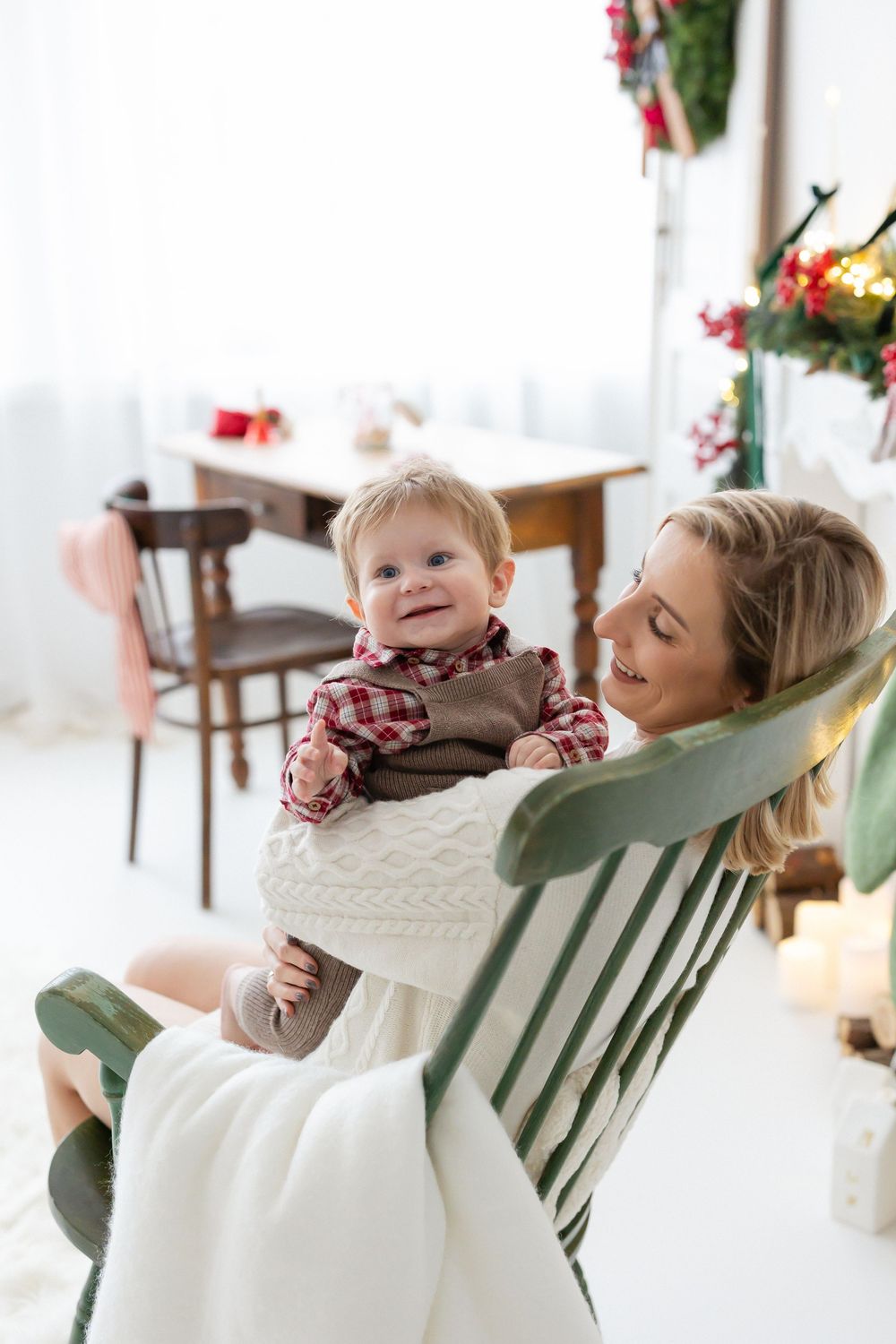 Smiling mother in a cream knit sweater holds her baby in a plaid shirt and vest on a green rocking chair near Christmas decorations.