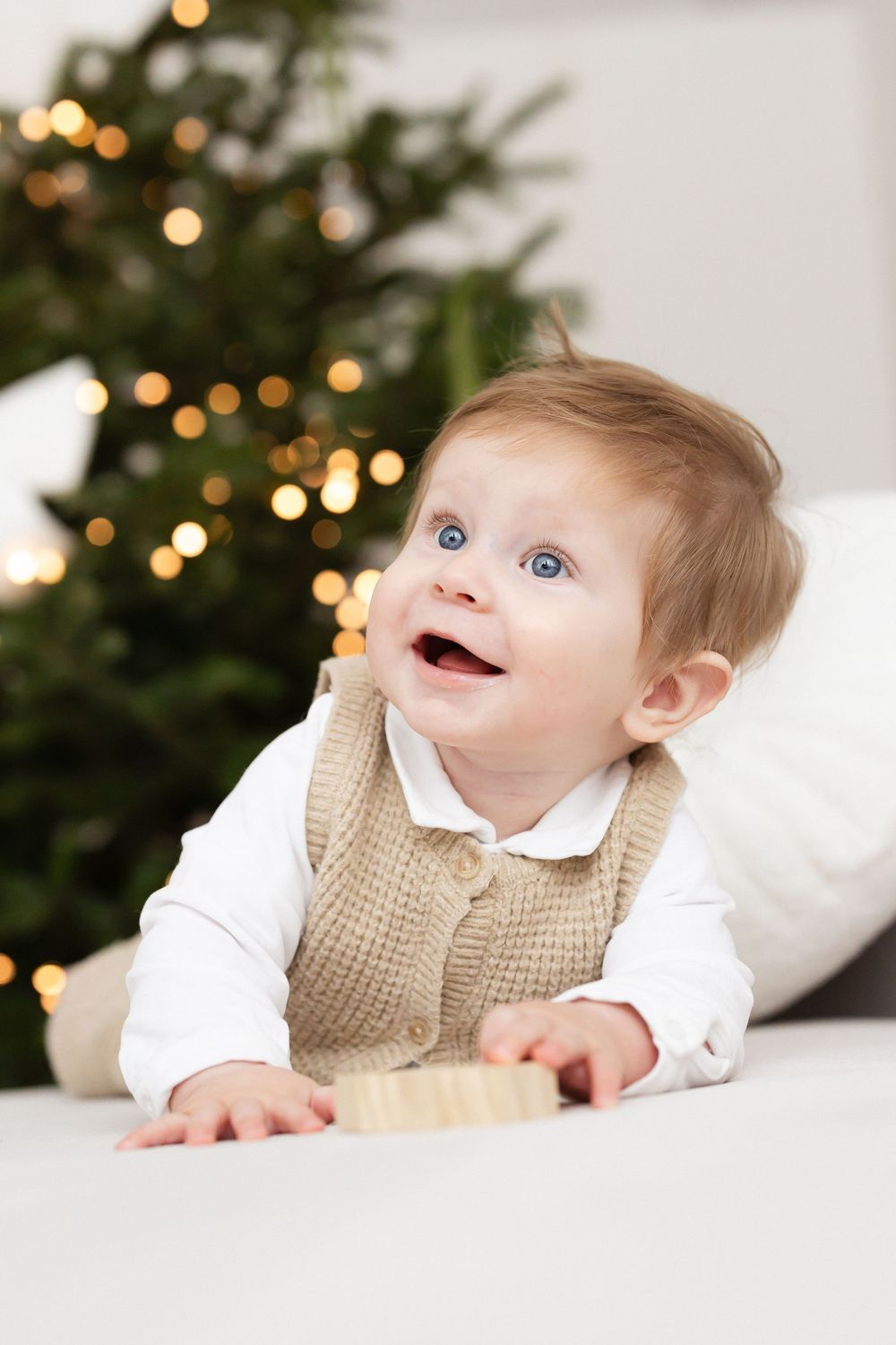 Smiling baby in a knit tan vest and white shirt crawls on a soft white surface beside a wooden block, with a glowing Christmas tree behind.