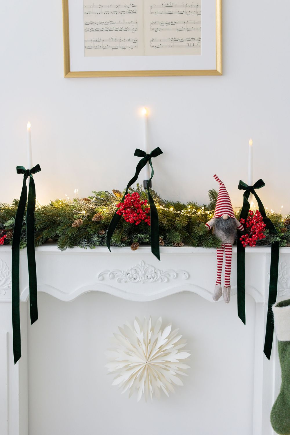 White mantel decorated with an evergreen garland, red berries, dark green velvet bows, a gnome ornament, candles and a paper star.