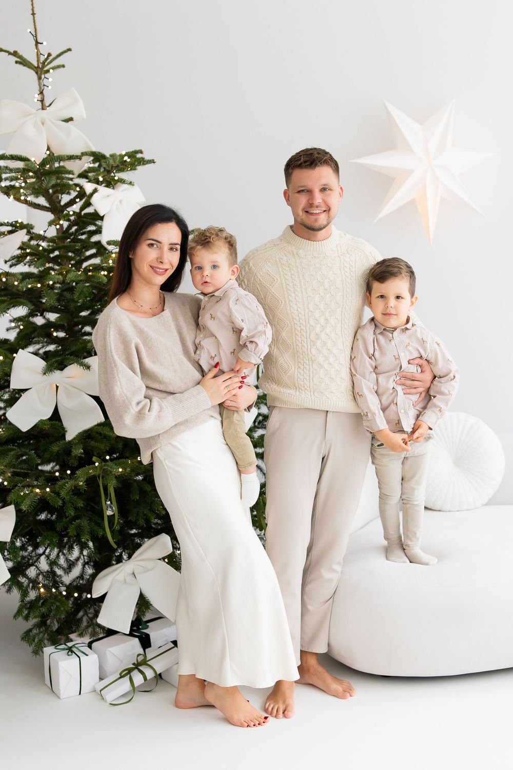 Smiling family of four in matching cream outfits poses barefoot beside a decorated Christmas tree with white bows, gifts and a star lamp.