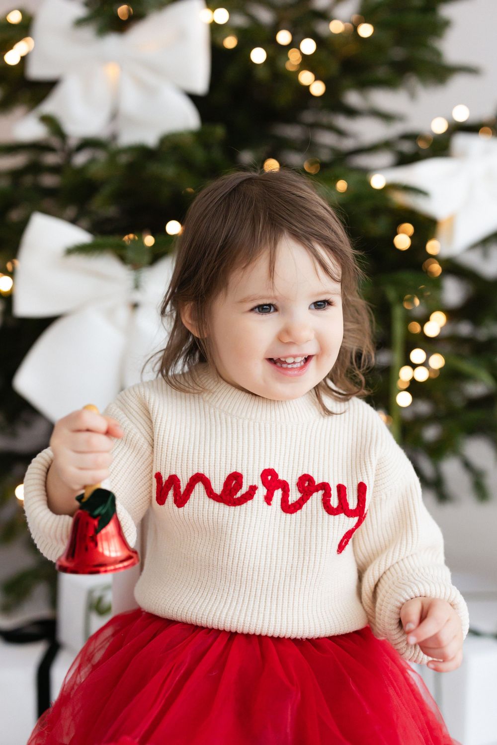 Smiling toddler in a cream knit "merry" sweater and red tulle skirt holds a red bell in front of a decorated Christmas tree.
