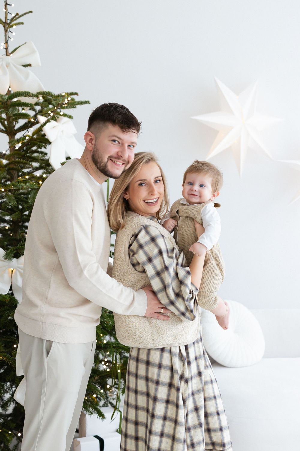 Smiling couple in neutral cozy outfits hold a laughing baby beside a decorated Christmas tree and white paper star lantern.