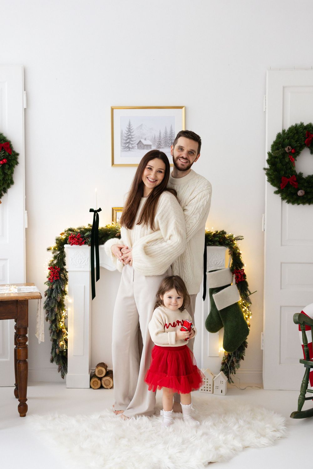 Smiling family in cozy cream outfits poses by a garland-draped fireplace with wreaths and a stocking, their toddler in a red tutu skirt.