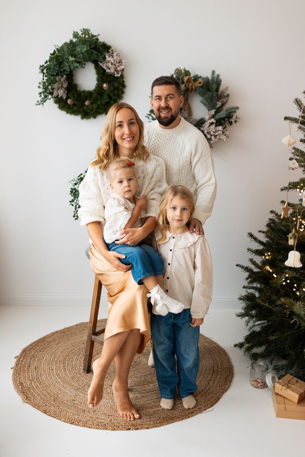 Smiling family of four poses together beside a decorated Christmas tree with wreaths on white wall.