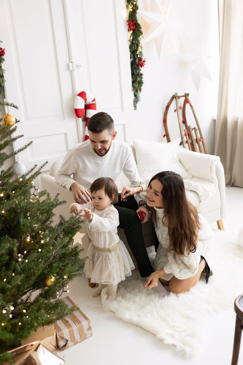Young family decorating a Christmas tree together, with toddler touching ornaments while parents watch and smile warmly.