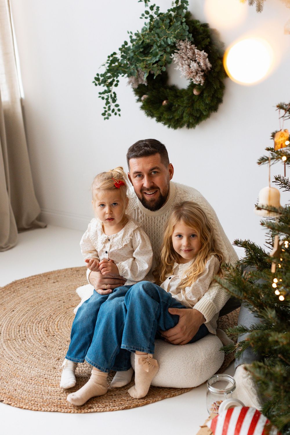 Smiling father sits on a rug with two young daughters beside a glowing Christmas tree indoors.