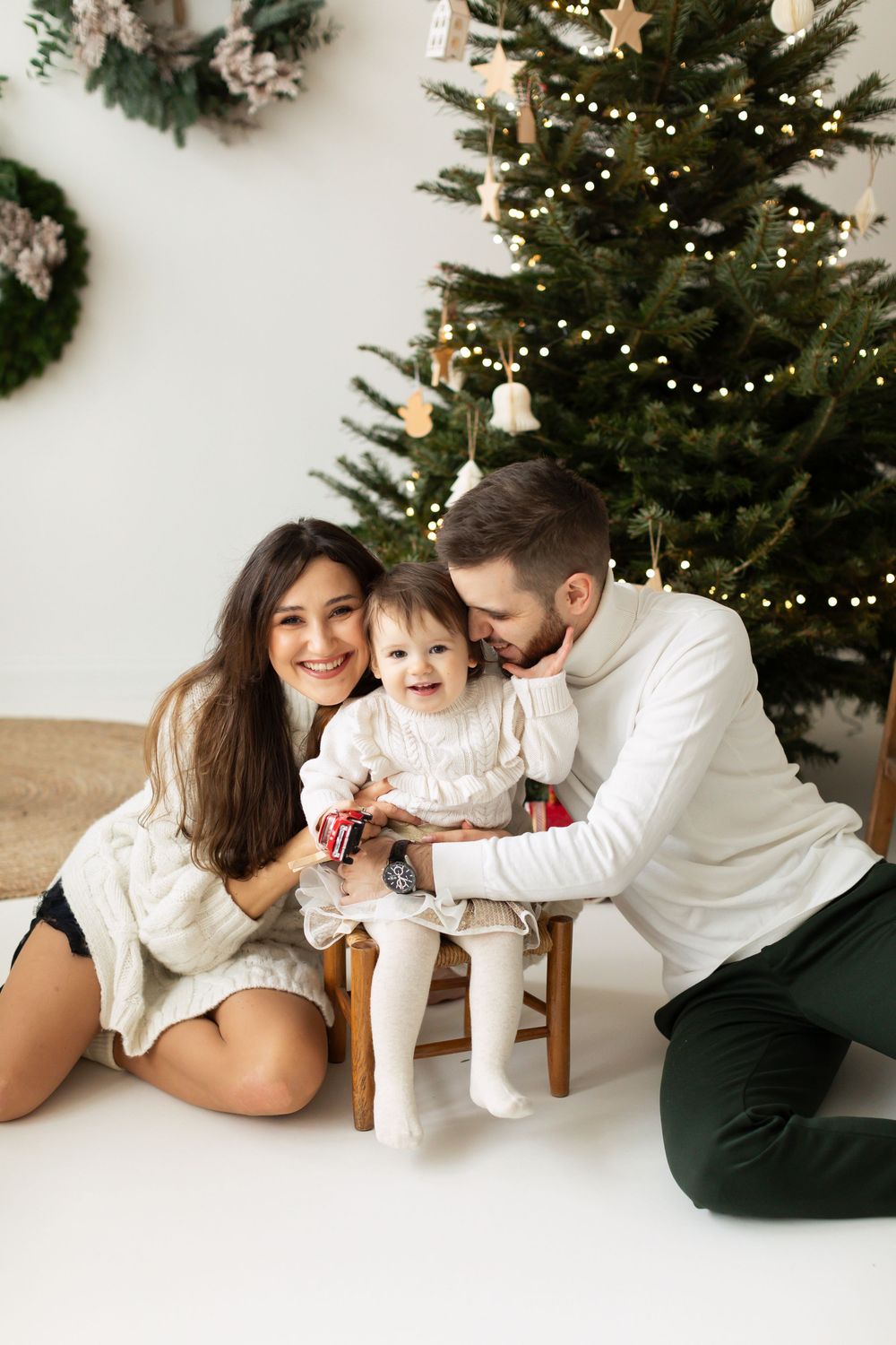 Happy young family with baby sitting near a decorated Christmas tree with warm glowing lights in a bright room.
