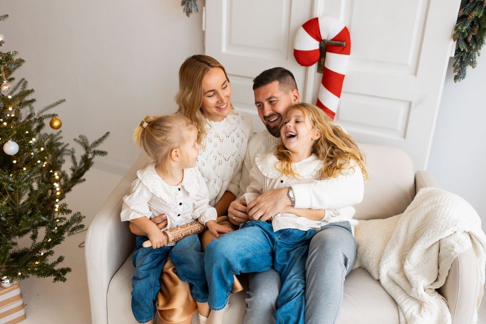 Joyful family of four laughing together on a sofa surrounded by Christmas tree and holiday decor.