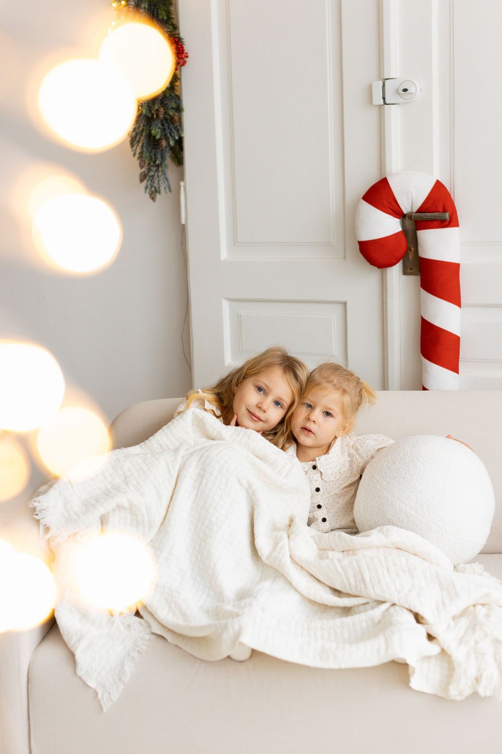 Two young siblings snuggle under a white blanket on a sofa with festive Christmas decorations in a bright white room.