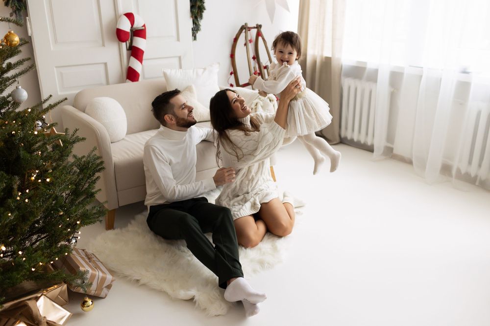 Joyful family playing together on the floor near a Christmas tree, parents lifting their baby girl in a white dress.