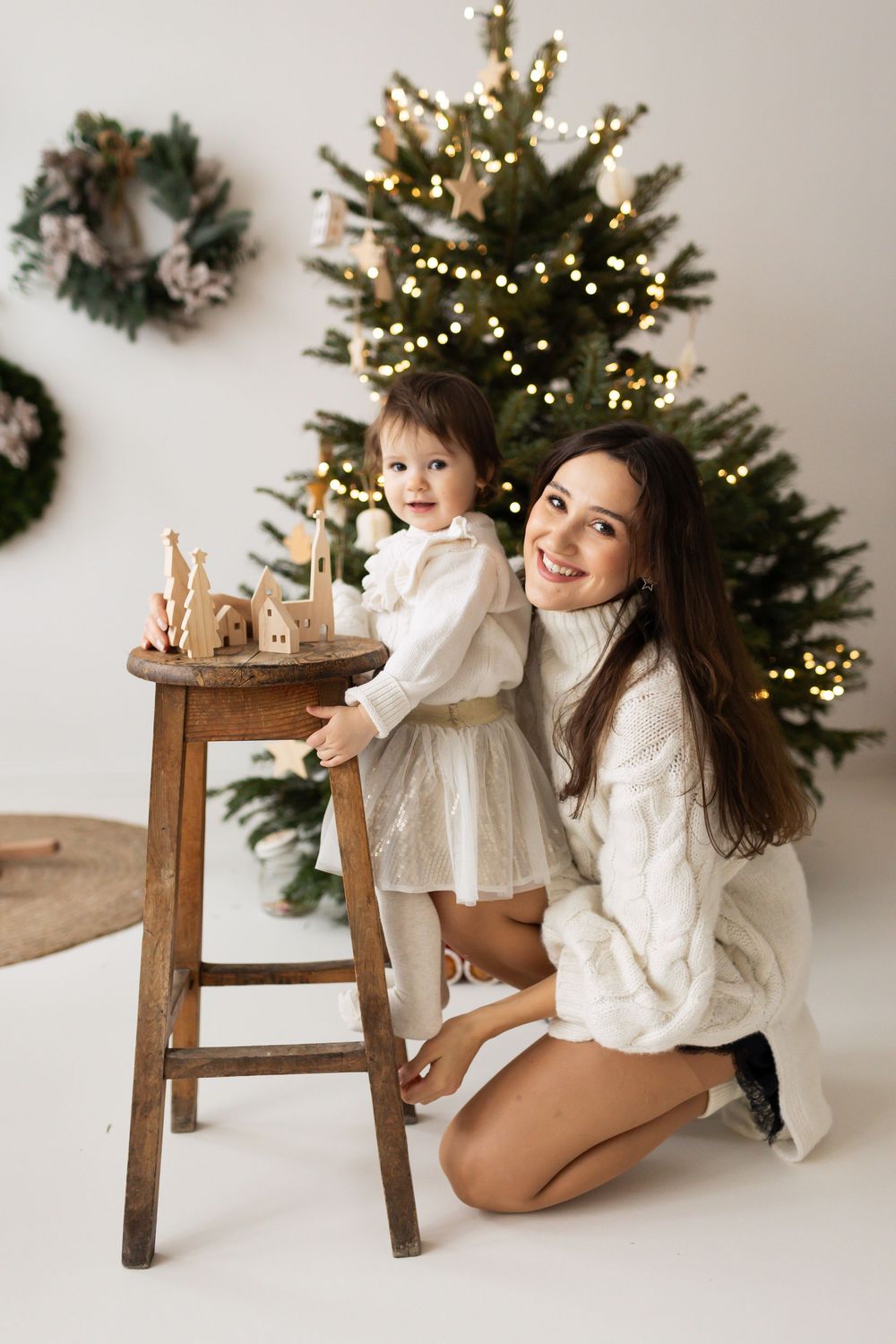 Smiling mother in white knit sweater kneeling beside toddler daughter standing at a wooden stool near Christmas tree.