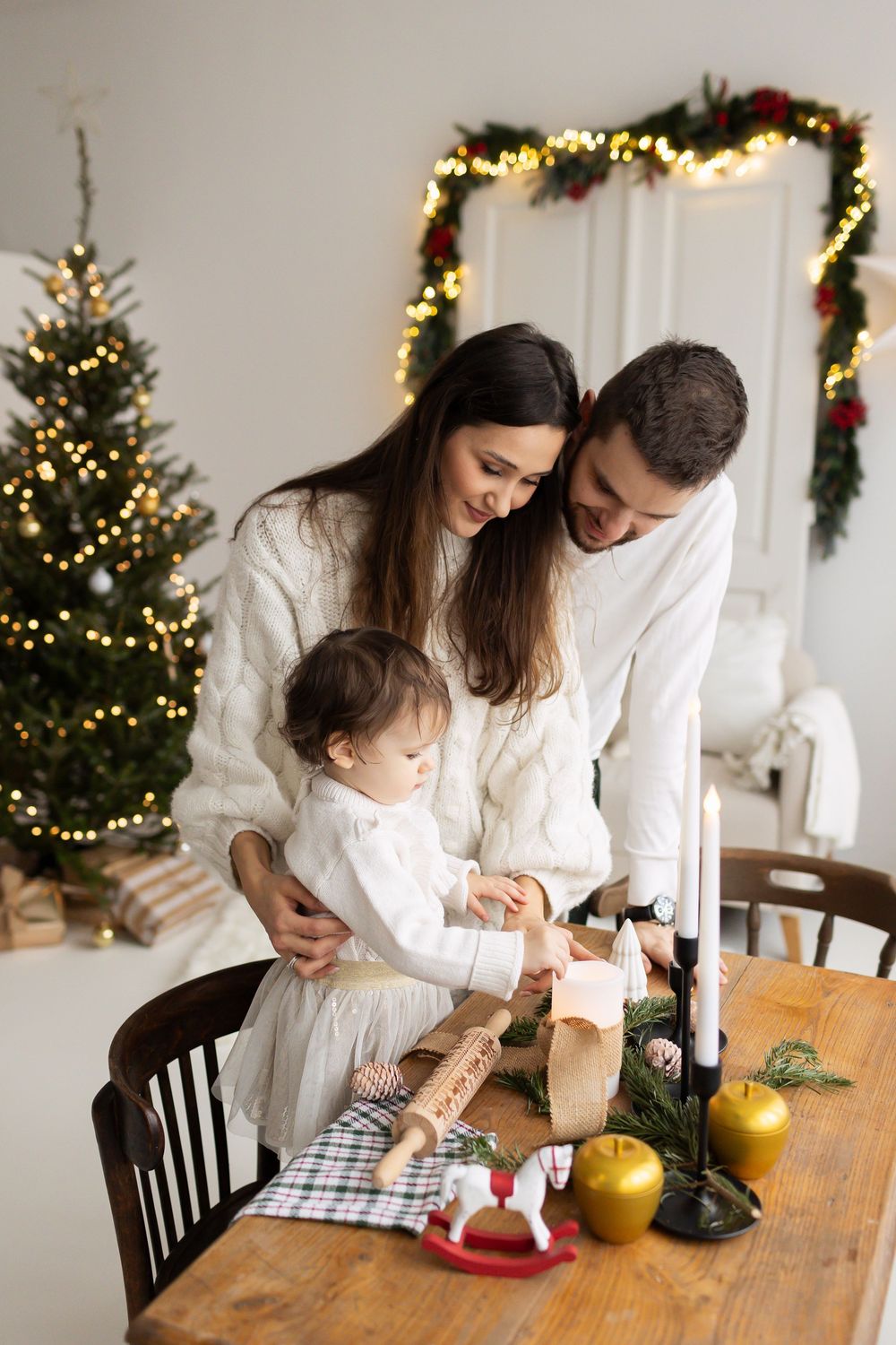 Young family in white sweaters gathered around a festive table with candles and Christmas decorations near a lit tree.