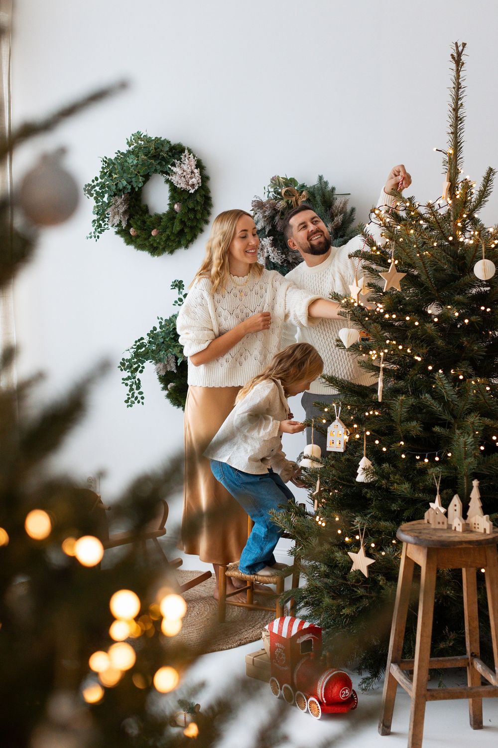 Happy family decorating a Christmas tree together in a bright white room with wreaths on the wall.