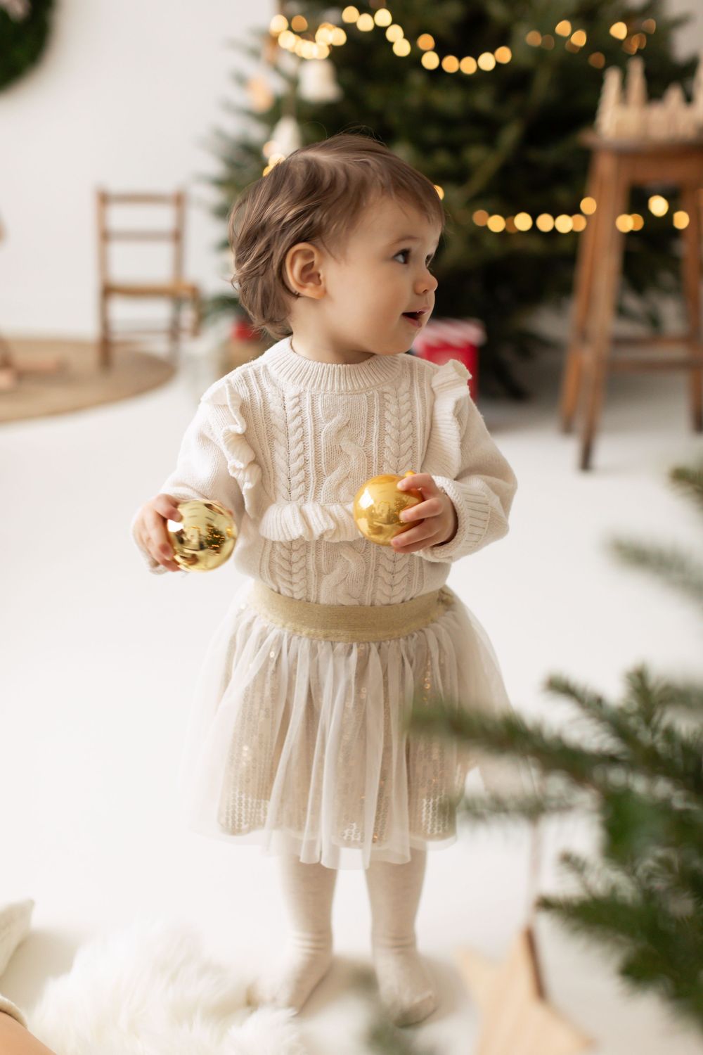 Adorable toddler girl in white knit sweater and tulle skirt holding gold Christmas ornaments near a decorated tree.