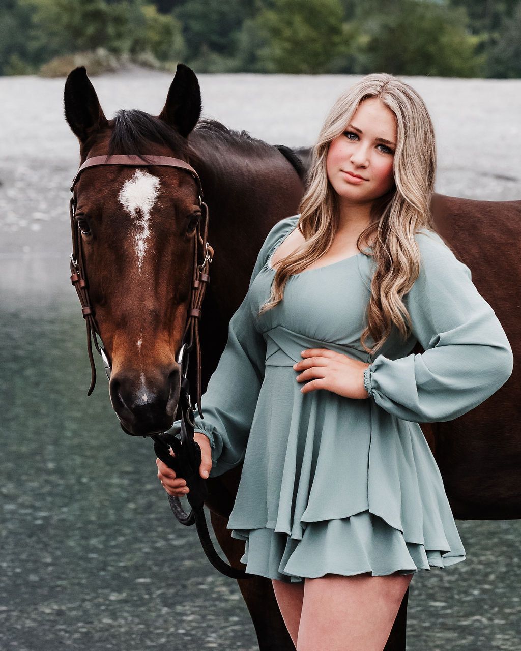 Senior photo session with girl in sage green dress poses elegantly with a dark brown horse beside a serene lake.