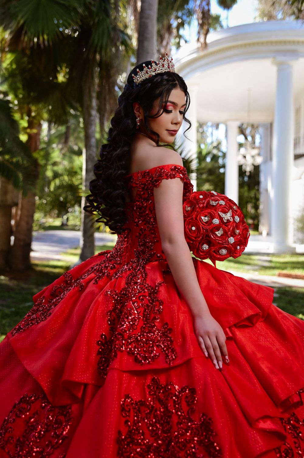 An ornate red ball gown with off-shoulder design and crystal embellishments photographed in a garden setting.