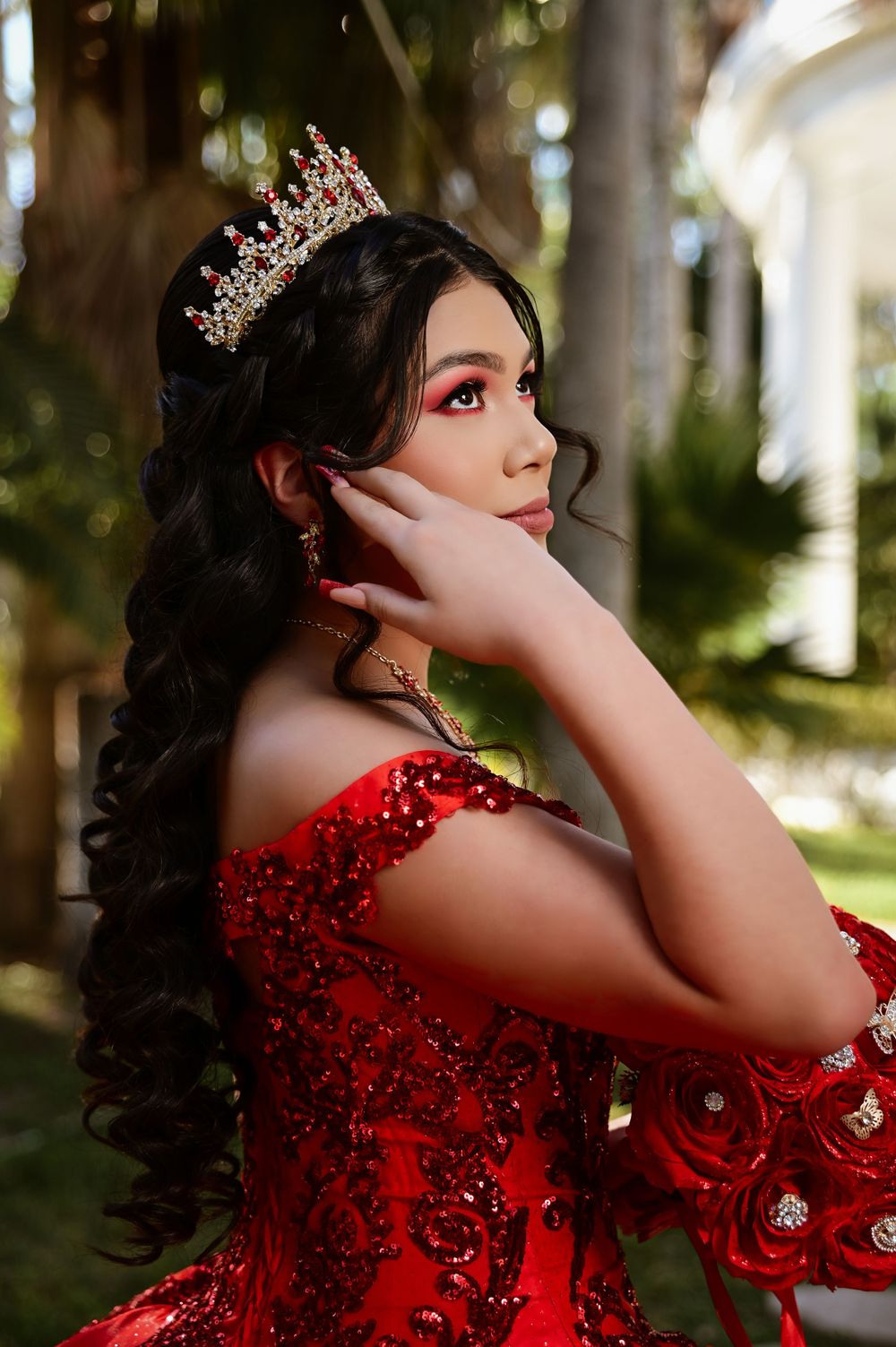 A jeweled crown adorns long dark curls against a backdrop of tropical palm trees.
