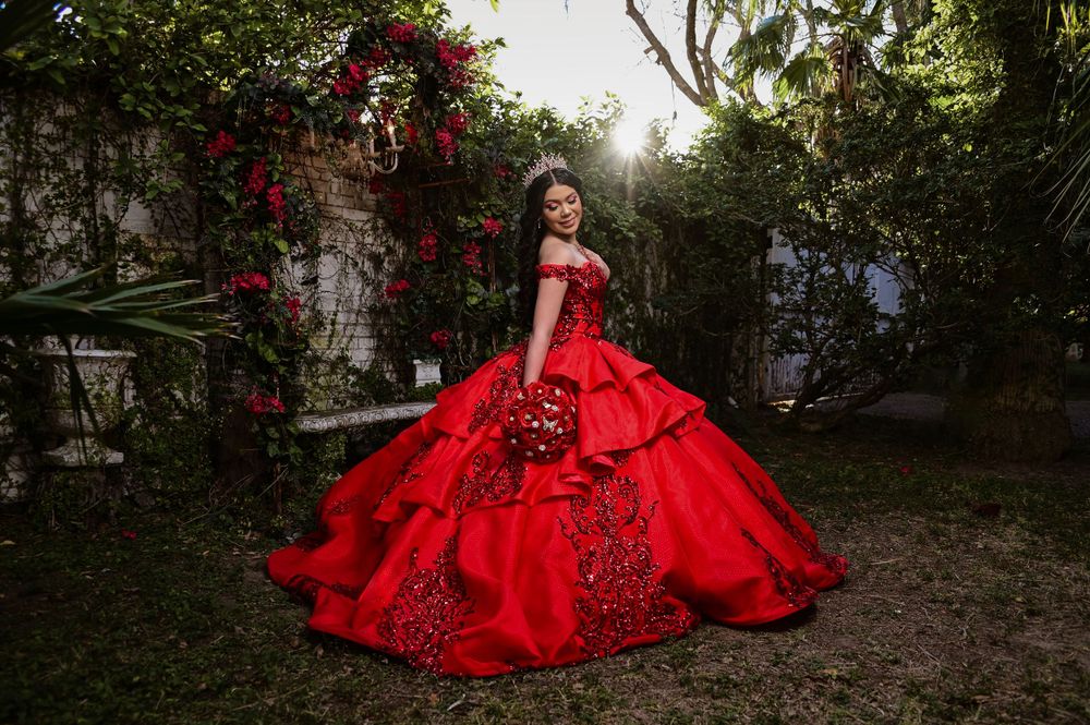 Vibrant red quinceañera dress photographed in a garden setting with climbing roses.