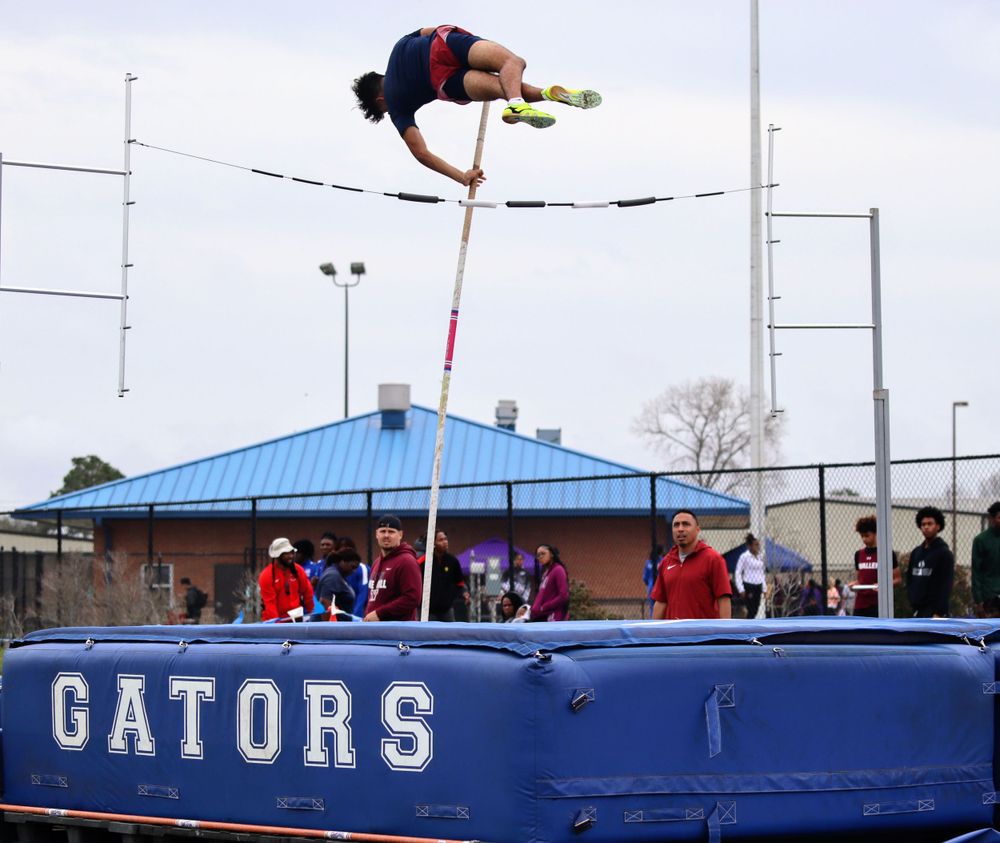 High school pole vaulter clears the bar at outdoor track meet with Gators branding visible.