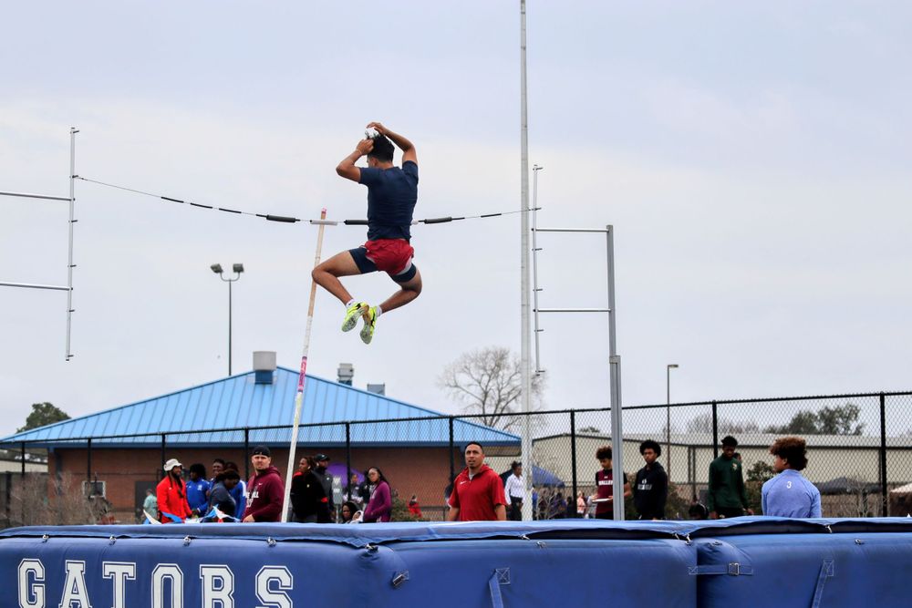 Athlete performs pole vault attempt at track competition with blue landing mat and Gators signage.