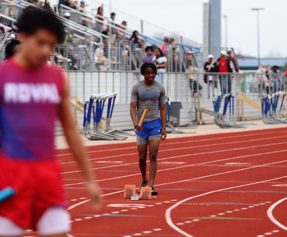 Track athletes running on a red track during a competitive race with spectators in the bleachers.