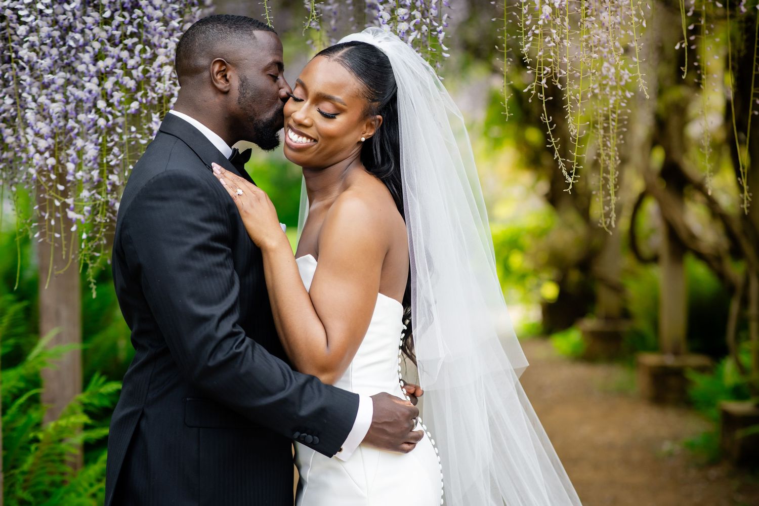 A couple shares a romantic moment under blooming wisteria in a garden setting.