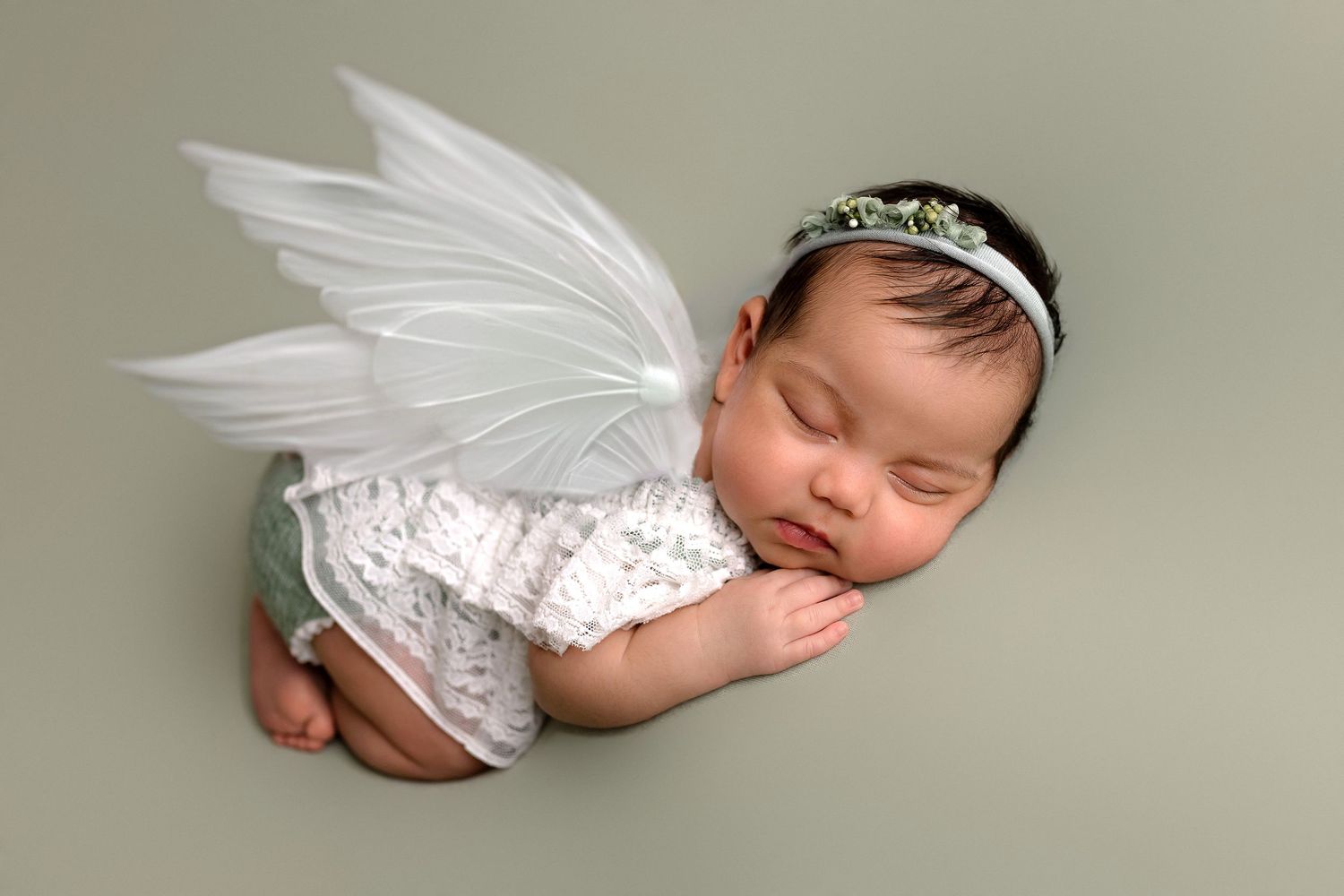 Sleeping newborn wearing white lace outfit and angel wings lies peacefully on grey backdrop in studio portrait.