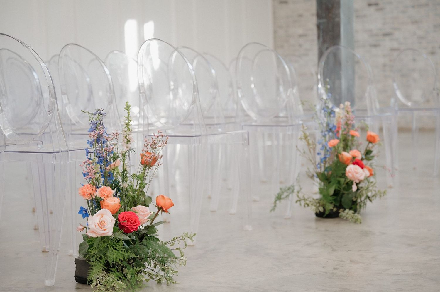 Clear ghost chairs line up for a wedding ceremony with colorful floral arrangements of coral and navy blooms beside each aisle.