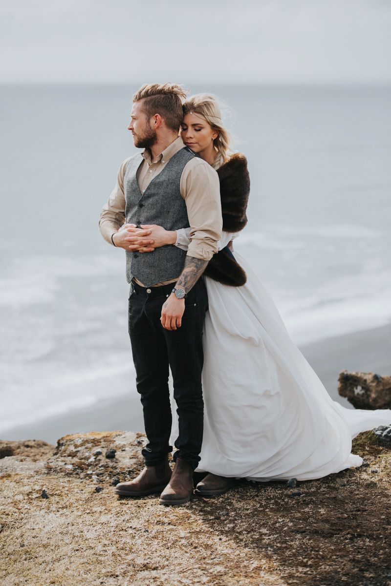 A romantic wedding couple embraces on a coastal cliff overlooking a misty ocean landscape.