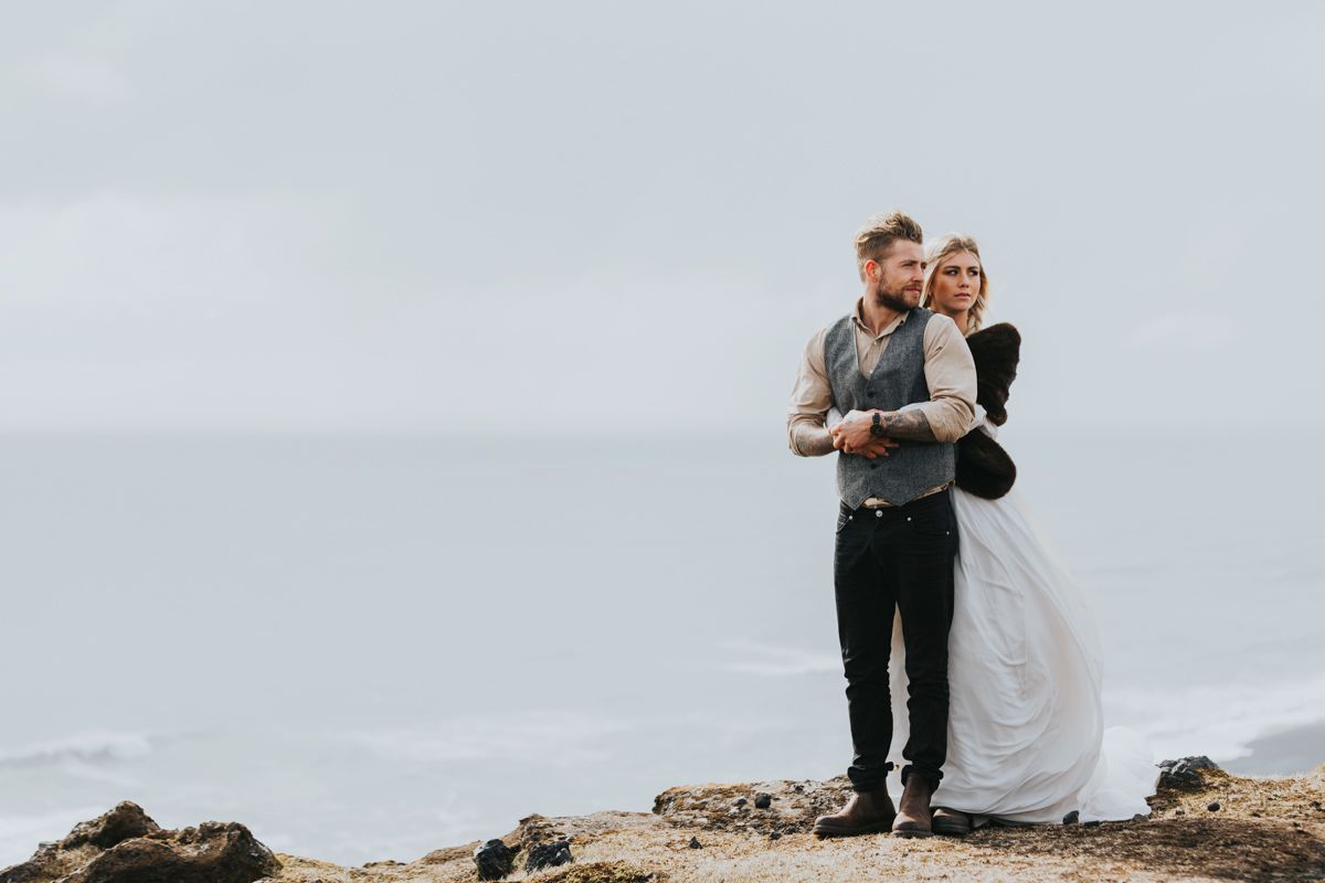 Newlyweds stand together on rocky oceanside cliff during moody overcast day.