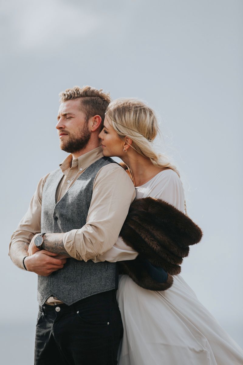 Intimate wedding portrait showing couple in rustic attire against cloudy coastal backdrop.