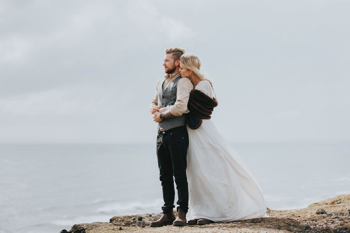 Wedding couple stands embracing on rocky ocean overlook during overcast day.