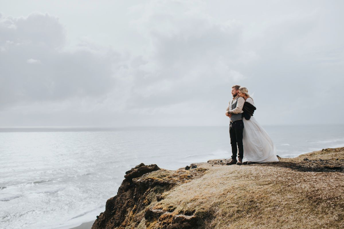 Dramatic wedding photo of couple standing on cliff edge overlooking stormy ocean.