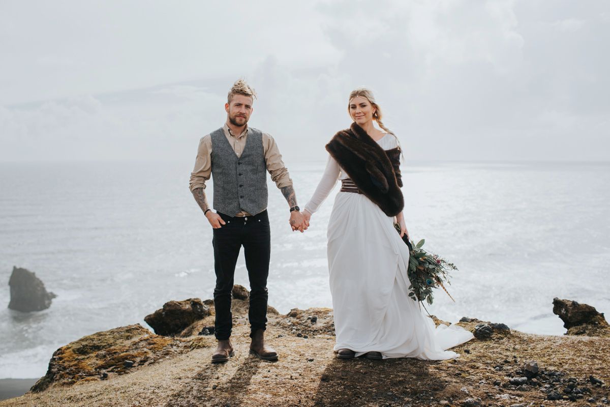 Modern wedding couple holds hands while standing on dramatic ocean cliff.
