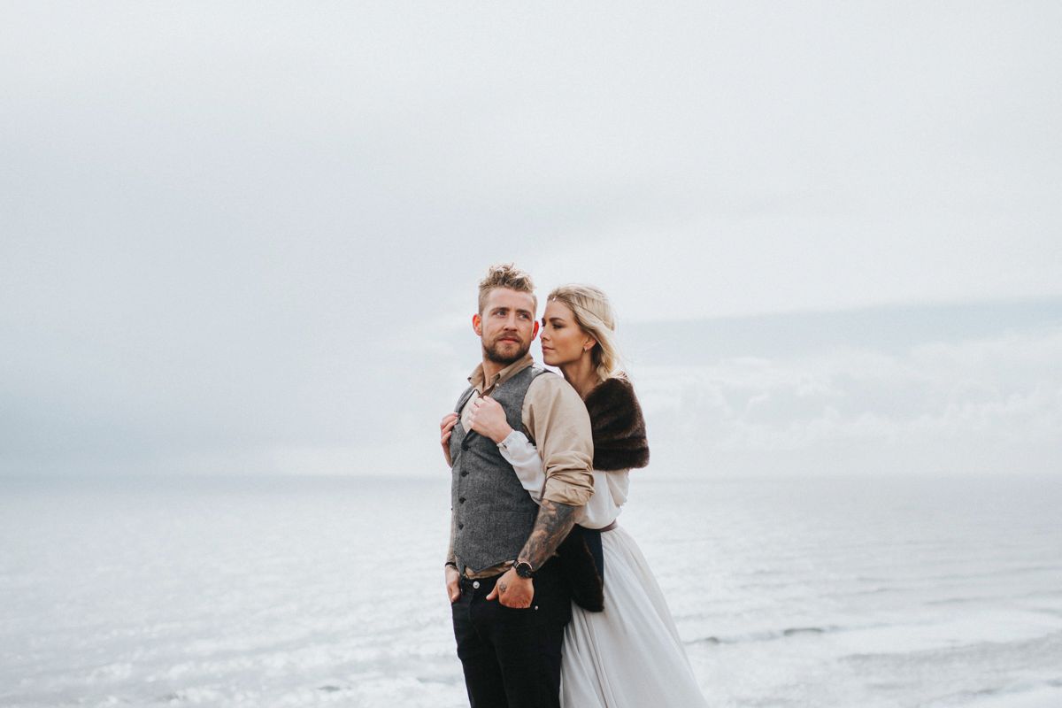 A couple in wedding attire poses against a foggy ocean backdrop on a rocky cliff.