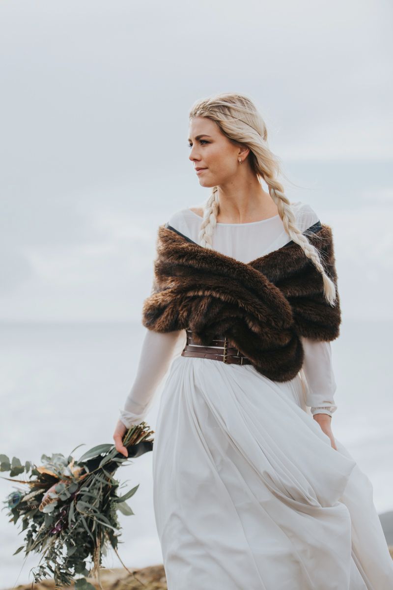 Bride in flowing white dress and fur wrap holds bouquet against moody coastal backdrop.