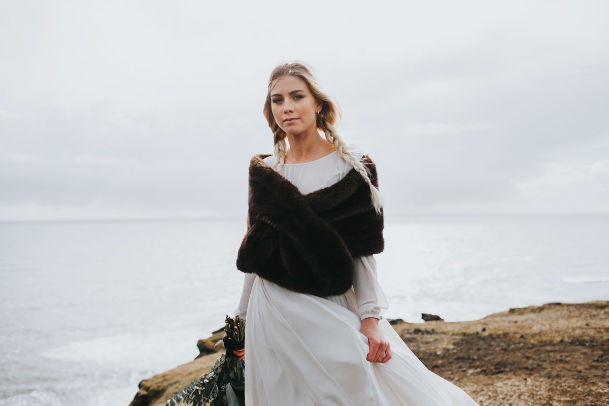 Bride in white dress and dark fur wrap stands on cliff edge holding rustic bouquet.